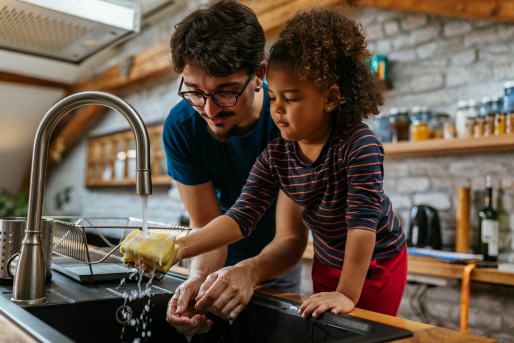 A man with glasses and a young girl washing dishes together in a kitchen sink, smiling as they wash dishes